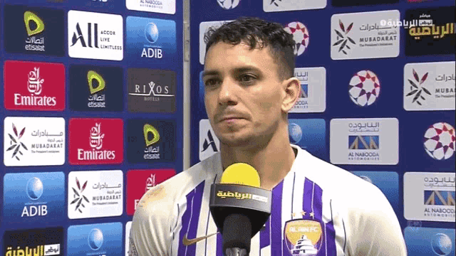 a man in a purple jersey is talking into a microphone in front of a wall of advertisements for emirates and adib