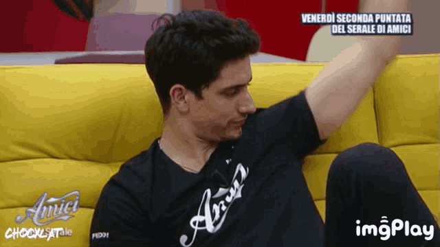 a man is sitting on a yellow couch wearing an amici t-shirt