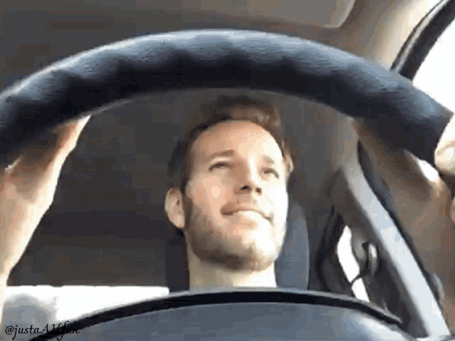 a man with a beard is driving a car with justa4life written on the bottom right