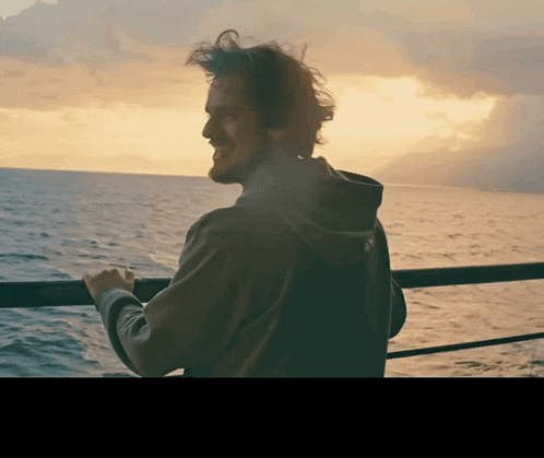 a man in a hoodie stands on a balcony overlooking the ocean at sunset