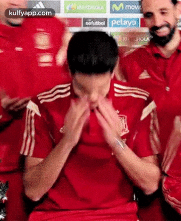 a man in a red shirt is covering his face with his hands while a group of men applaud .