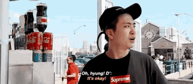 a man wearing a hat and a supreme shirt is standing in front of a coca cola machine .