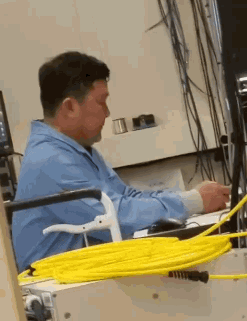 a man in a blue shirt sits at a desk with a yellow cord