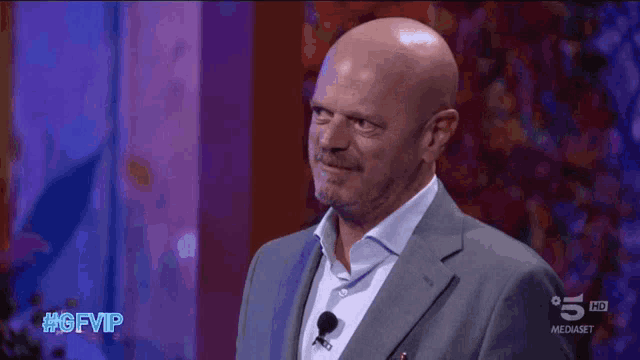 a bald man in a suit stands in front of a purple wall with #gfvip written on it