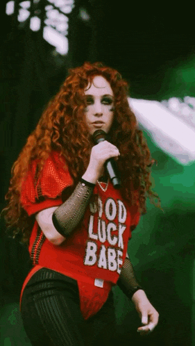 a woman singing into a microphone wearing a red shirt that says god luck babe