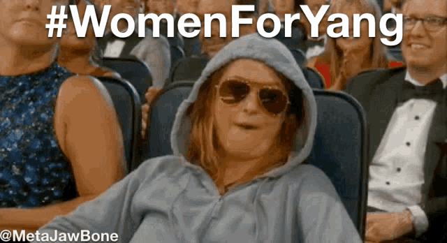 a woman wearing a hoodie and sunglasses sits in a crowd of people with the hashtag #womenforyang