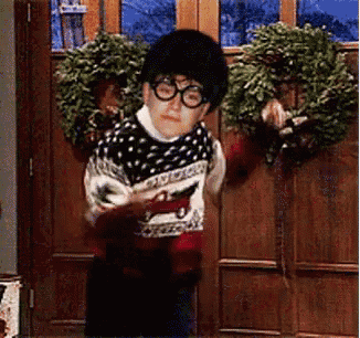 a young boy wearing glasses and a sweater is standing in front of a door with a wreath hanging on it .