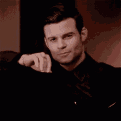 a man in a black suit and tie is sitting in a chair with his hand on his shoulder .