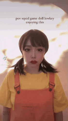 a girl in a yellow shirt and orange overalls is playing a game called squid game doll lowkey