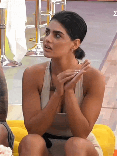 a woman in a white tank top is sitting on a yellow couch with her hands folded