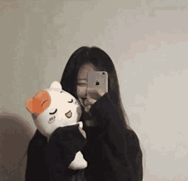 a woman is taking a selfie with her phone while holding a stuffed cat .
