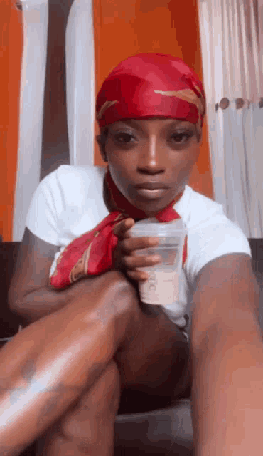 a woman wearing a red head scarf is sitting on a couch holding a plastic cup .