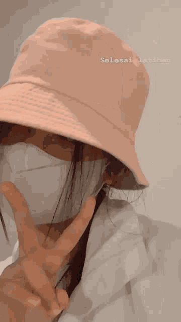 a woman wearing a pink hat and a white face mask shows a peace sign
