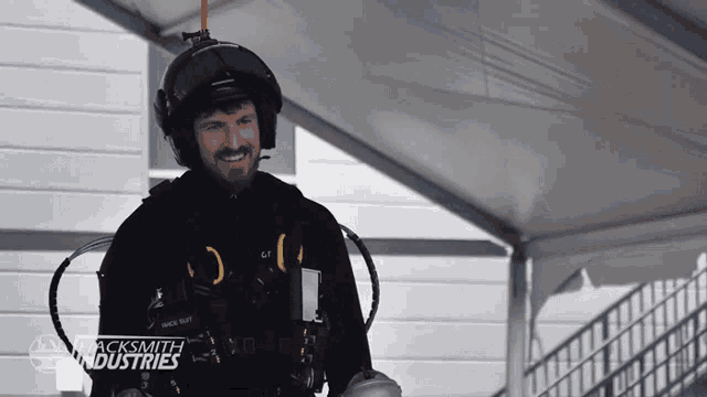 a man wearing a helmet and gloves is smiling in front of a sign that says acksmith industries