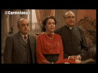a man in a suit stands next to a woman in a red dress and a priest in a black robe