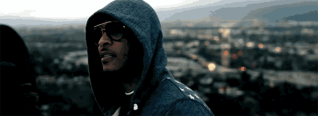 a man wearing a hoodie and sunglasses stands in front of a city at night