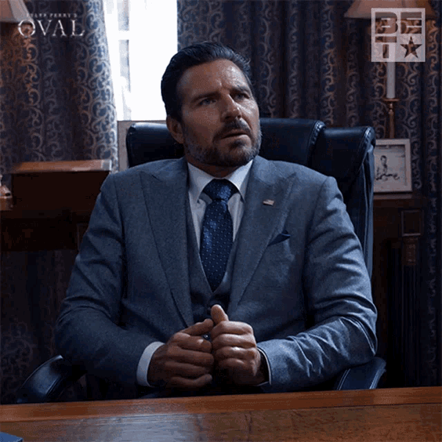 a man in a suit and tie is sitting at a desk in front of a sign that says " the oval "