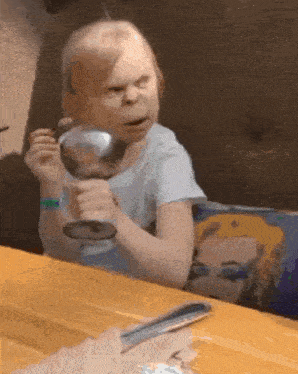 a little girl is sitting at a table holding a glass of wine and making a funny face .