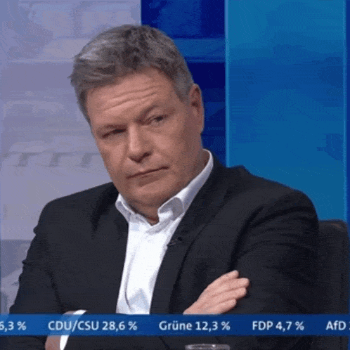 a man in a suit sits with his arms crossed in front of a screen that says grüne