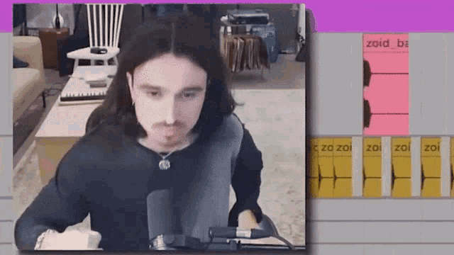a man with long hair is sitting in front of a microphone and a screen that says zaid ba