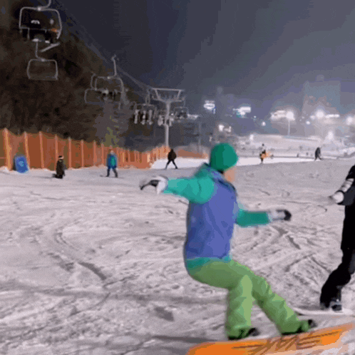 a snowboarder wearing a blue jacket and green pants rides down a snowy slope at night