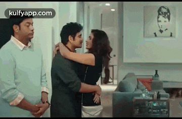 a man and a woman are hugging in a living room while a man stands behind them .