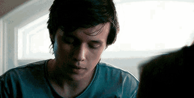 a young man in a blue shirt is looking down at something while sitting in front of a window .