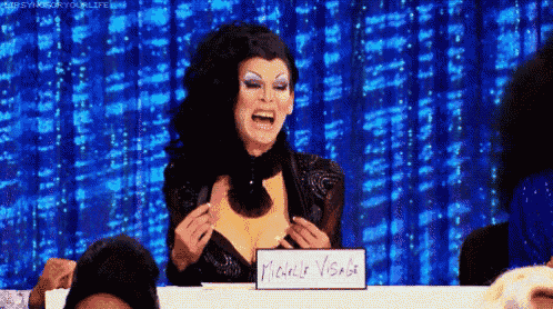 a drag queen is sitting at a table holding a sign that says michelle visage .