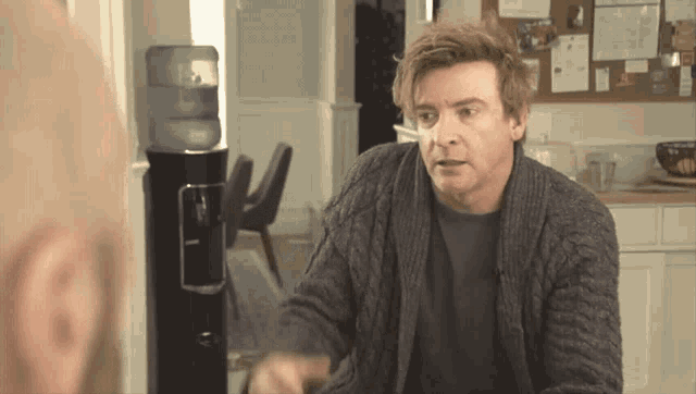 a man in a grey sweater is standing in front of a water cooler