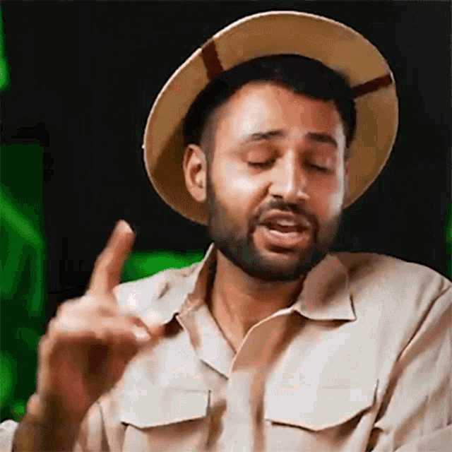 a man with a beard wearing a hat and khaki shirt is making a gesture with his hand .