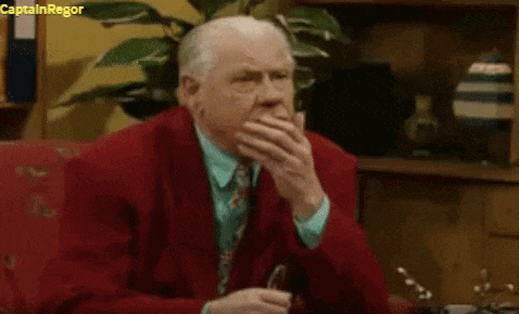 a man in a red suit and tie is sitting on a couch covering his face with his hand .