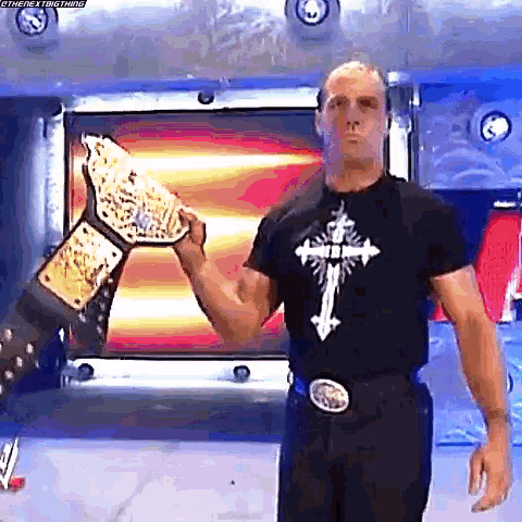 a man in a black shirt with a cross on it holds a wrestling championship belt