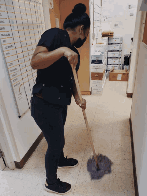 a woman wearing a mask is mopping the floor in a hallway with a list of names on the wall behind her