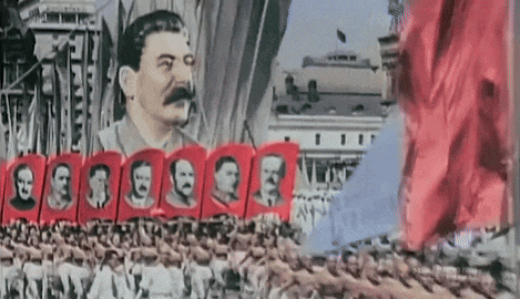 a man with a mustache stands in front of a group of people holding red flags with their faces on them