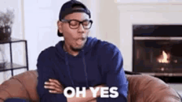 a man wearing glasses and a baseball cap is sitting on a couch with his arms crossed and says oh yes .