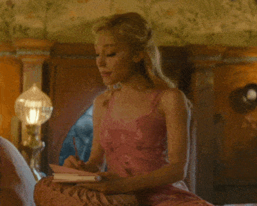 a woman in a pink dress sits on a bed writing in her notebook