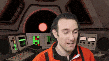 a man in an orange jacket is standing in front of a control panel with a red star in the background