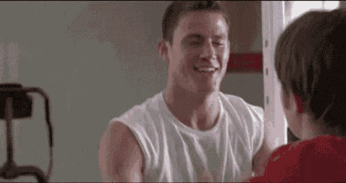 a man in a white tank top is smiling while looking at another man in a gym .