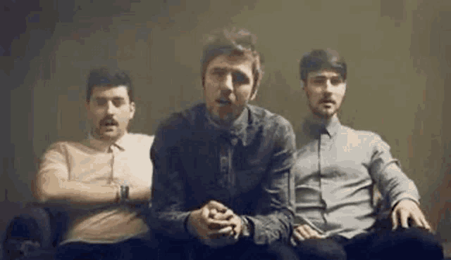 three men are sitting on a couch with their arms crossed and talking to the camera .
