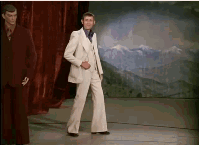 a man in a white suit is standing in front of a mountain backdrop