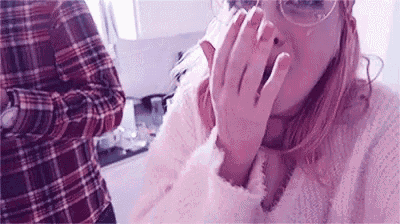 a woman wearing glasses is covering her mouth with her hands while a man in a plaid shirt stands behind her .