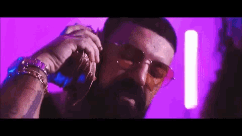 a man with a beard and sunglasses is talking on a cell phone in a purple room .
