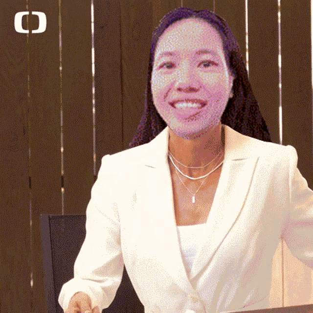 a woman wearing a white jacket and necklaces smiles in front of a wooden fence