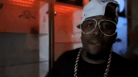 a man wearing a hat , sunglasses and a chain is standing in a dark room .