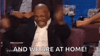 mike tyson is sitting on a couch with his arms in the air and says `` and we are at home '' .