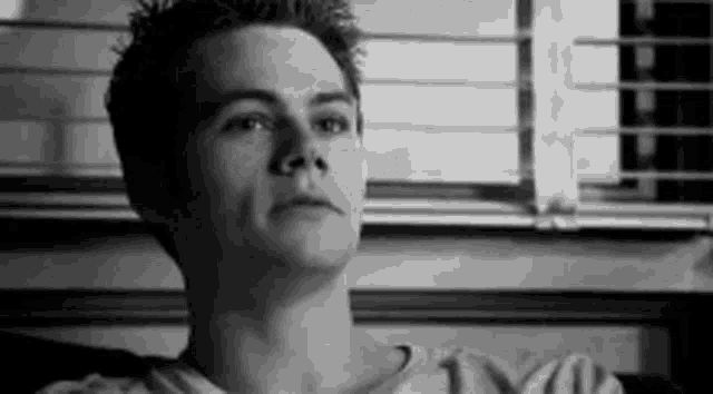 a black and white photo of a young man sitting in front of a window with blinds .