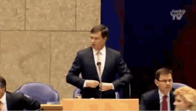 a man in a suit stands at a podium giving a speech in front of a lucky tv screen
