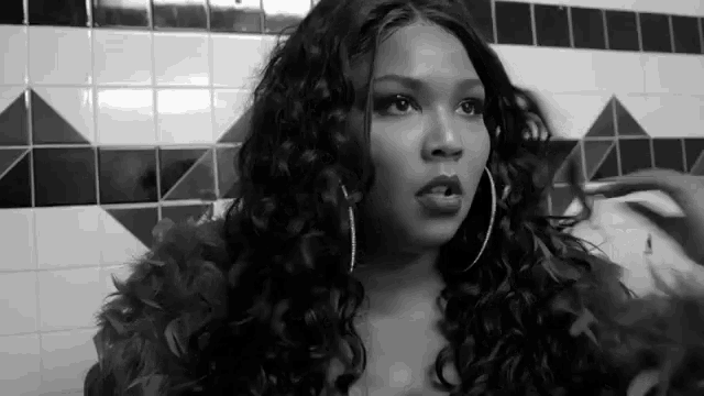 a black and white photo of a woman with long curly hair and hoop earrings standing in front of a tiled wall .