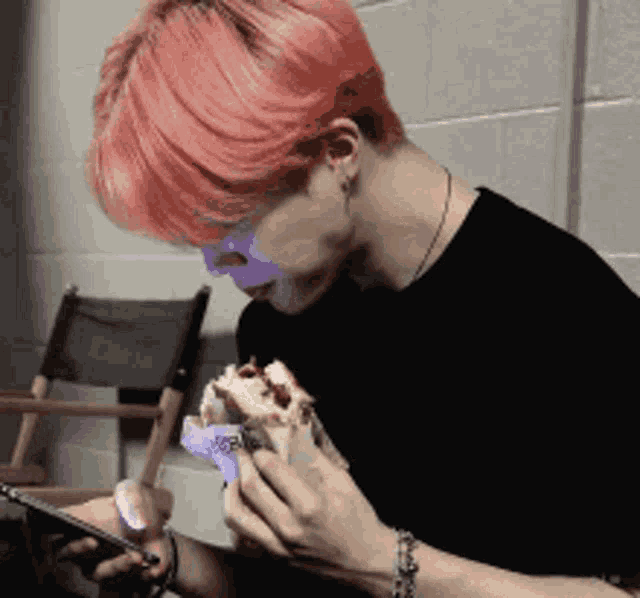 a man with pink hair is eating a sandwich while looking at his phone