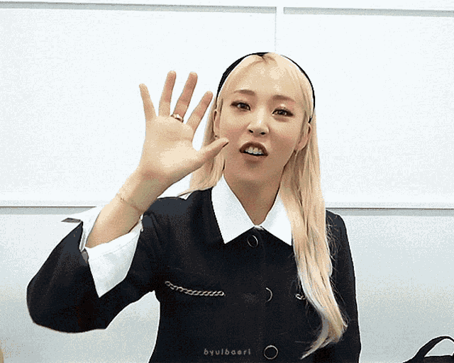 a woman wearing a headband and a black jacket waves her hand in front of a white wall